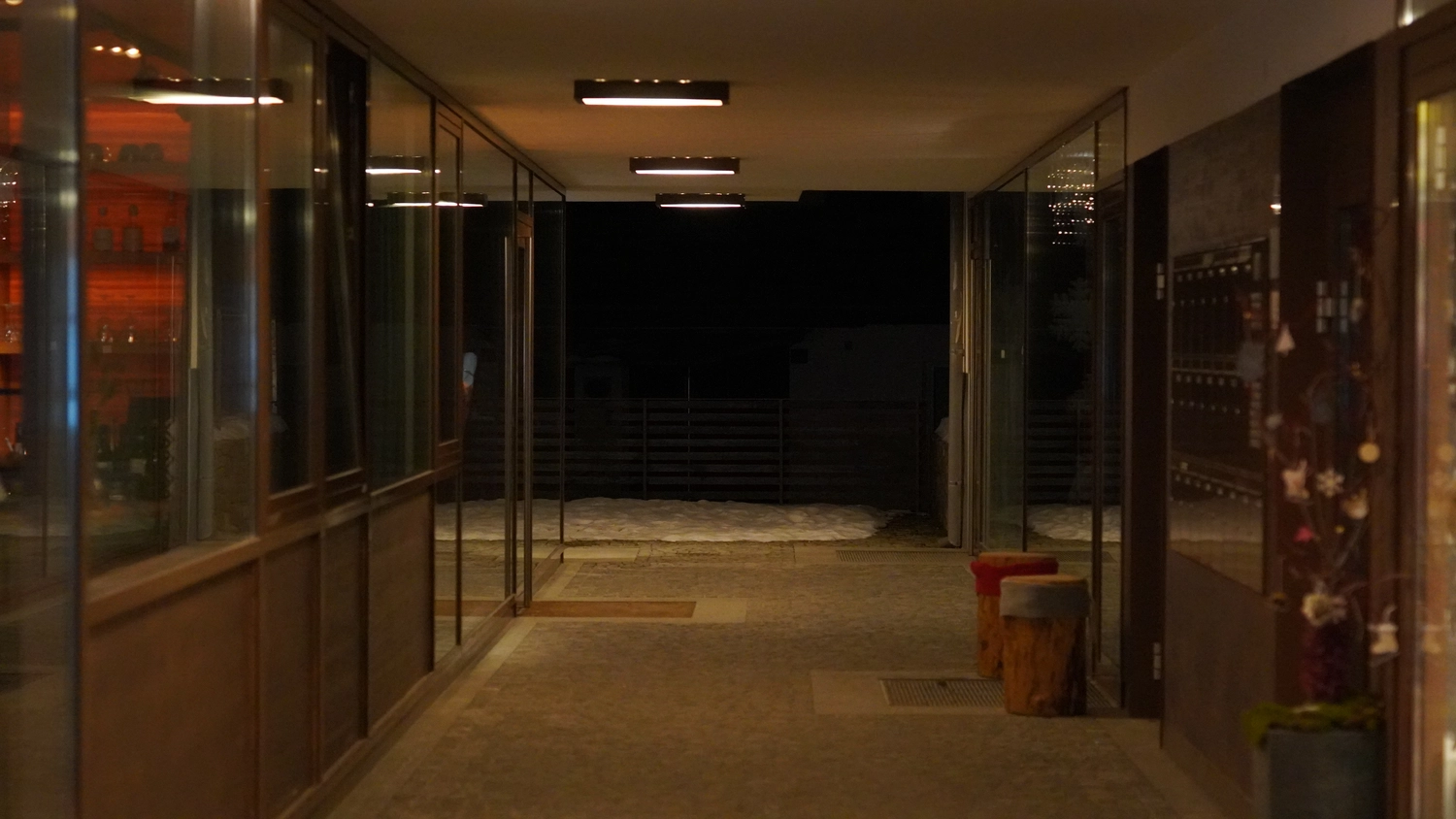 Sipho Studio - Drone visuel - Warmer Gang mit Glasfronten und Holzhockern führt hinaus in eine verschneite Nacht.