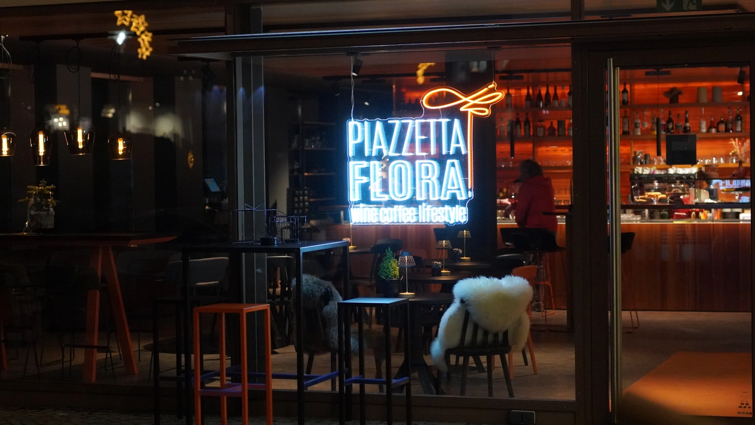 Sipho Studio - Drone visuel - Schaufenster mit leuchtend blau-orangefarbener Neonreklame „Piazzetta Flora“ über gemütlichen Sitzgelegenheiten.