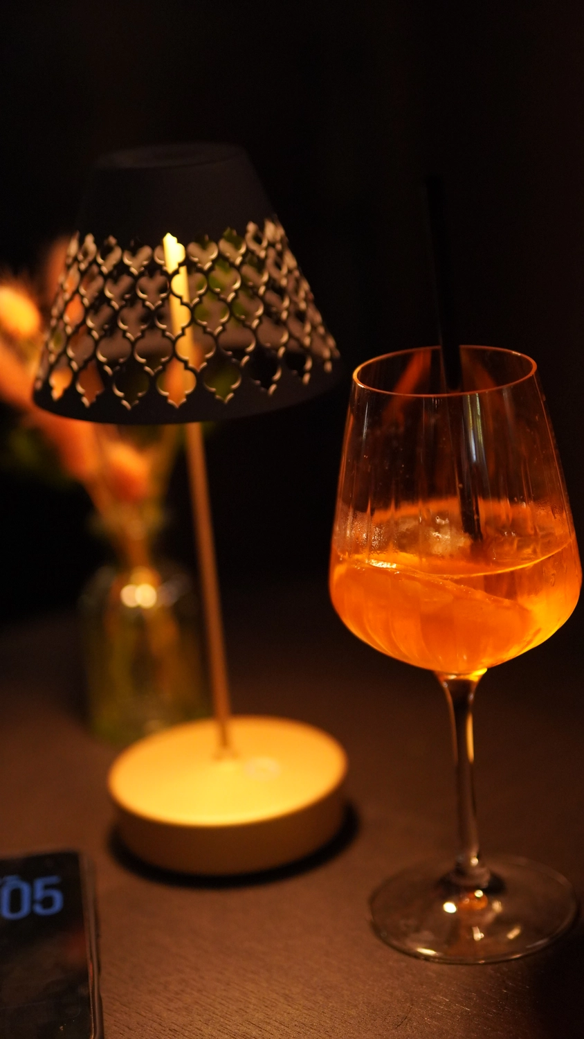 Sipho Studio - Drone visuel - Ein Orangencocktail in einem Weinglas steht neben einer kleinen, leuchtenden Tischlampe mit dekorativem Lampenschirm.