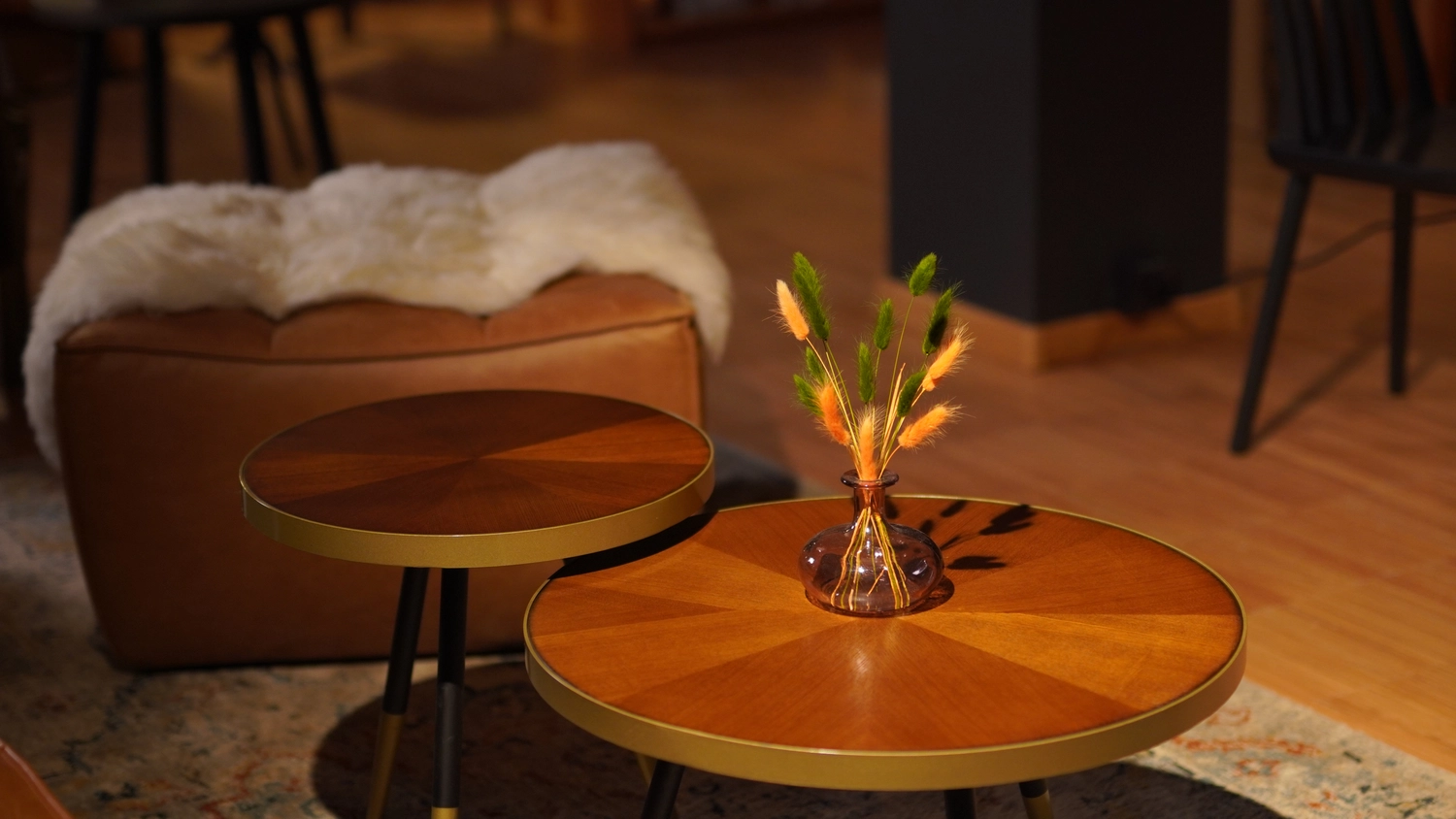 Sipho Studio - Drone visuel - Ein leuchtend orange-grünes Blumenarrangement in einer kleinen bernsteinfarbenen Glasvase steht auf einem runden Beistelltisch aus Holz.
