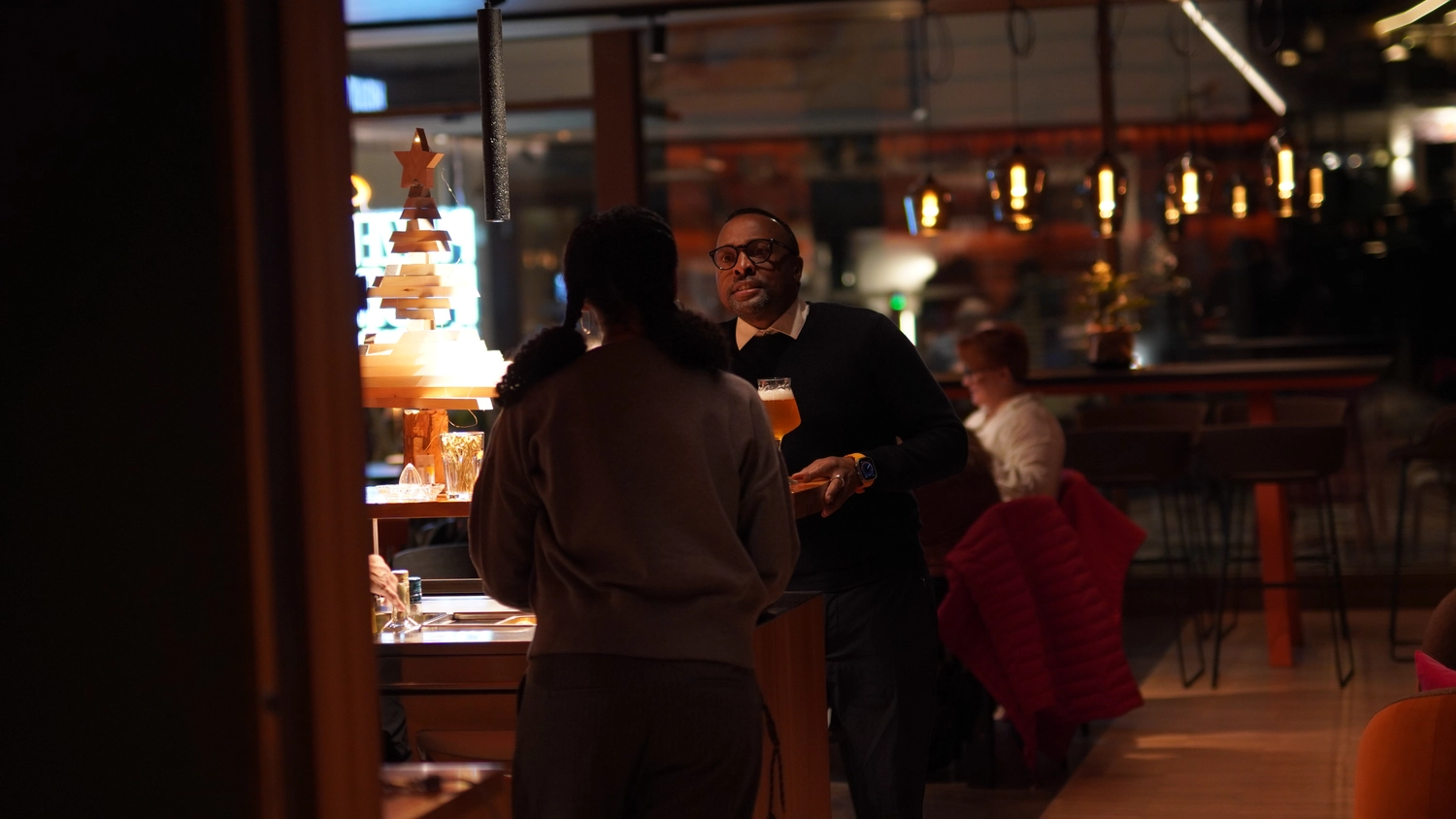 Sipho Studio - Drone visuel - Kellner mit Bier und Glas spricht an der Bar mit einer Frau in warm beleuchtetem Restaurant.