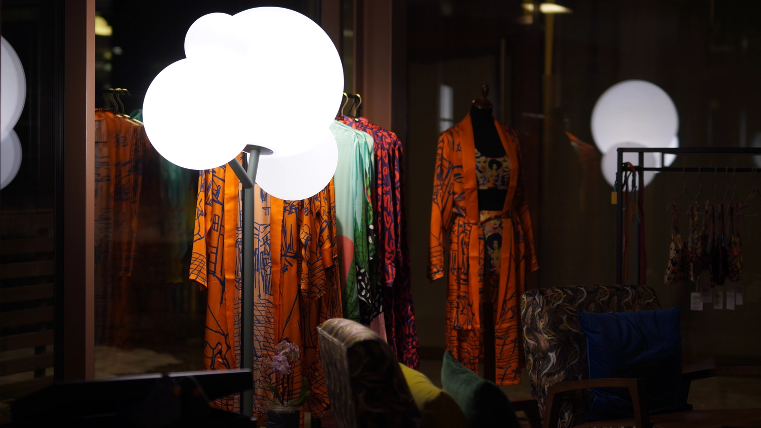 Sipho Studio - Drone visuel - Orange-grüne Seidenbademäntel mit leuchtenden Mustern hängen in einer schwach beleuchteten Boutique an einer Kleiderstange.