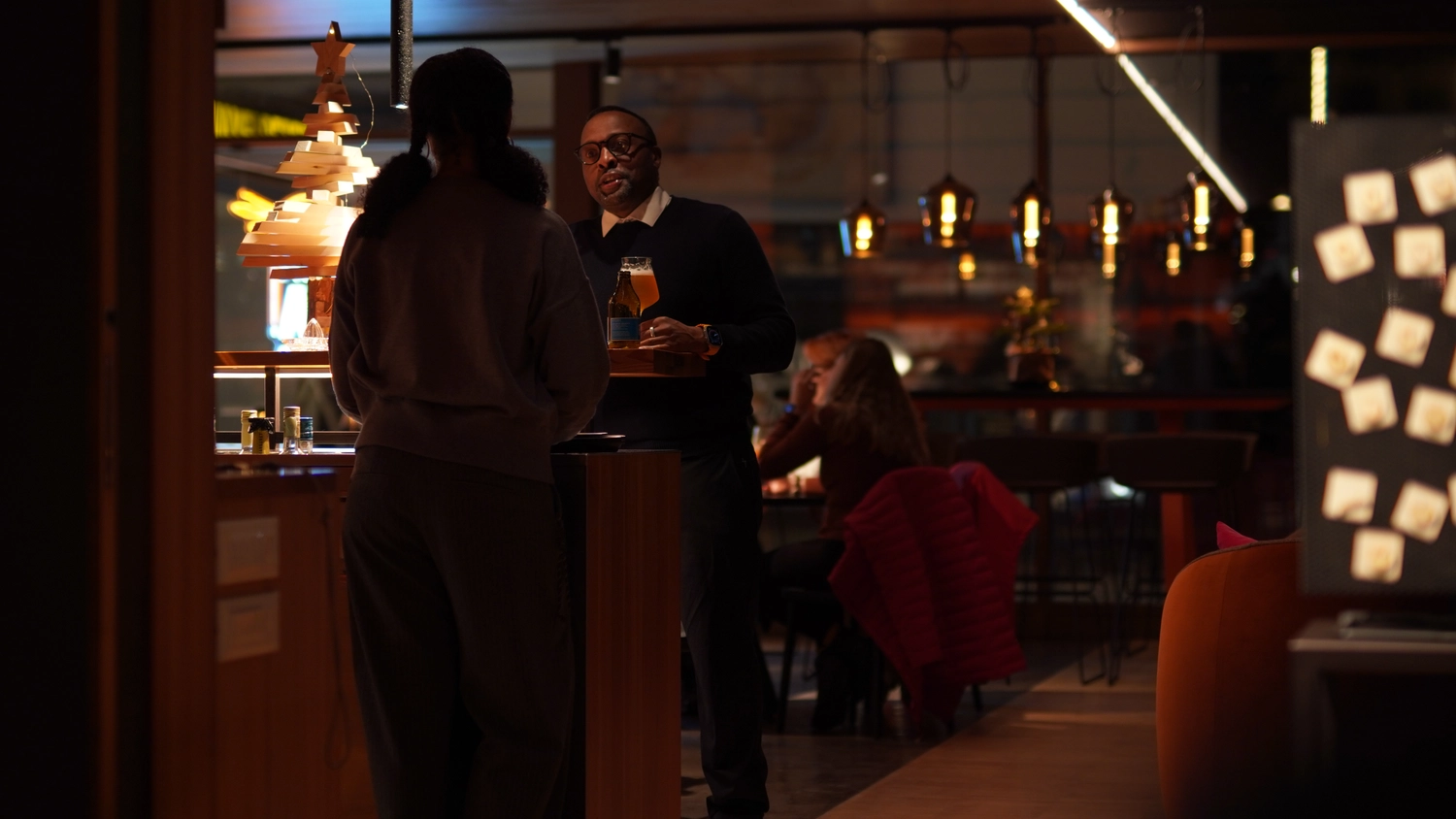 Sipho Studio - Drone visuel - Kellner mit Bier und Glas spricht an der Bar mit einer Frau in warm beleuchtetem Restaurant.