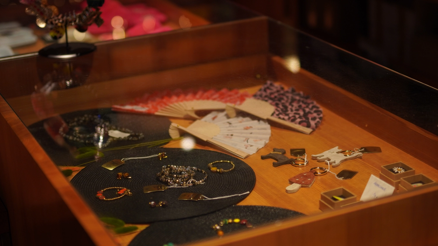 Sipho Studio - Drone visuel - Eine Sammlung kunsthandwerklichen Schmucks und Accessoires, angeordnet auf einer dunklen Ausstellungsmatte mit traditionellen faltbaren Handfächern im Hintergrund.