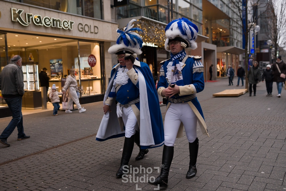 Sipho Studio - Drone visuel - Zwei Männer in blauen Uniformen und Federhüten laufen durch eine belebte Einkaufsstraße.