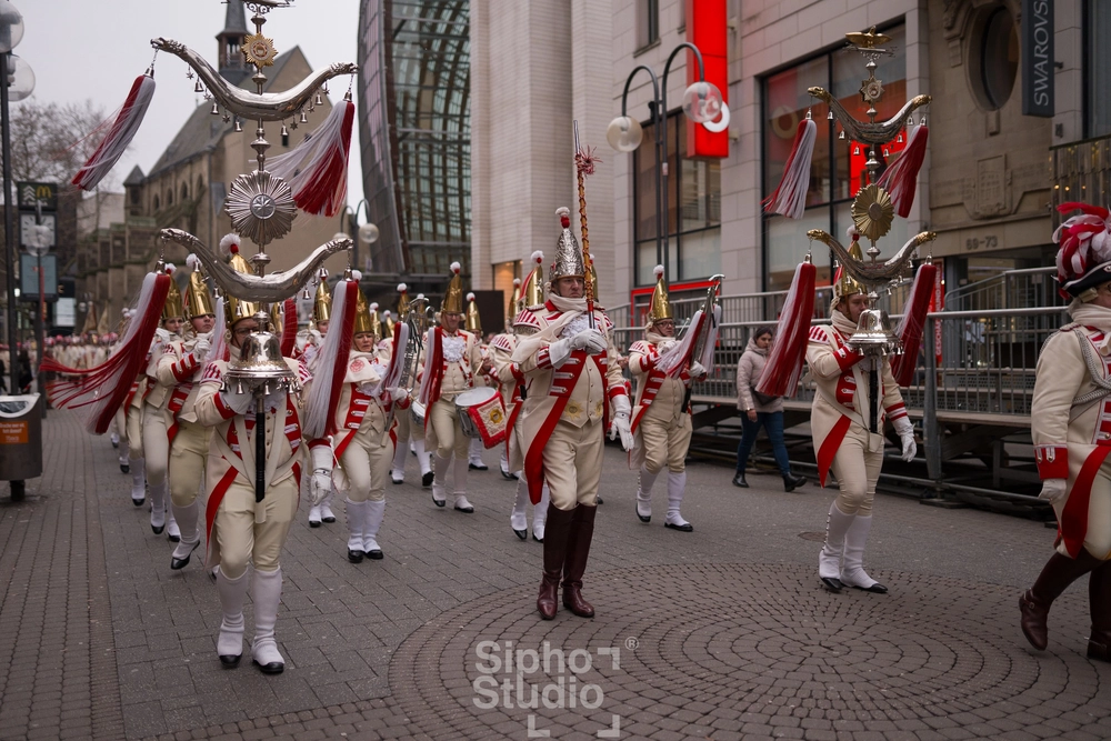 Sipho Studio - Drone visuel - Musikgruppe in roten und weißen Uniformen marschiert mit goldenen Helmen und Fanfaren durch die Stadt.
