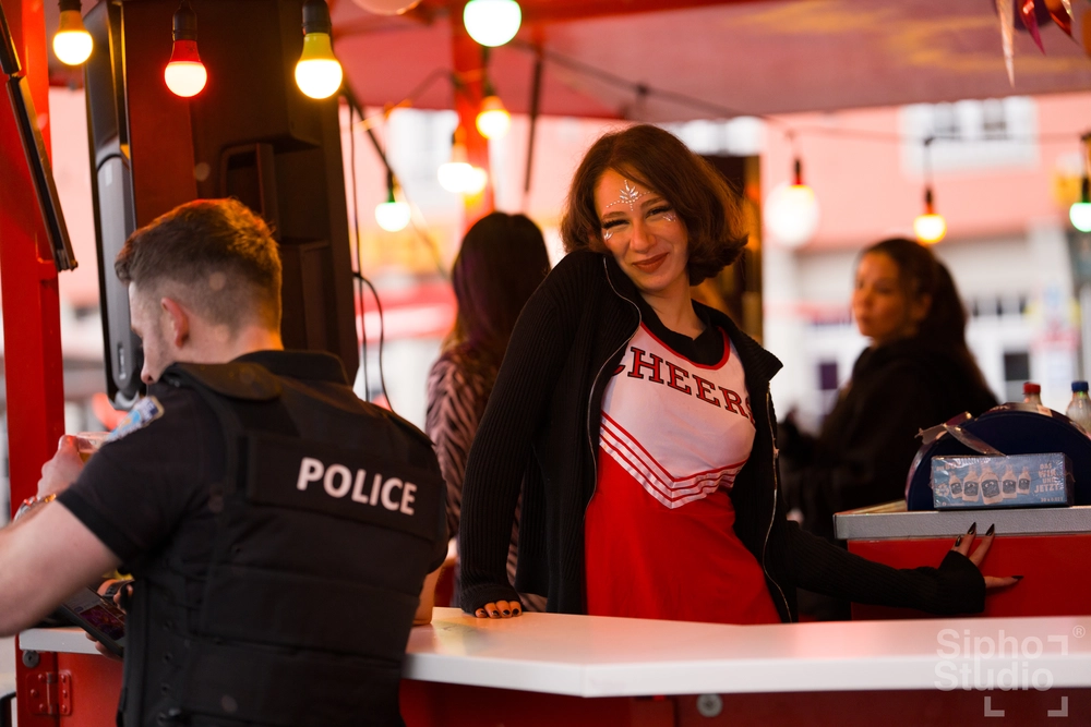 Sipho Studio - Drone visuel - Frau im roten Cheerleader-Shirt mit "CHEER" steht lächelnd an einem Tresen, daneben ein Polizist.