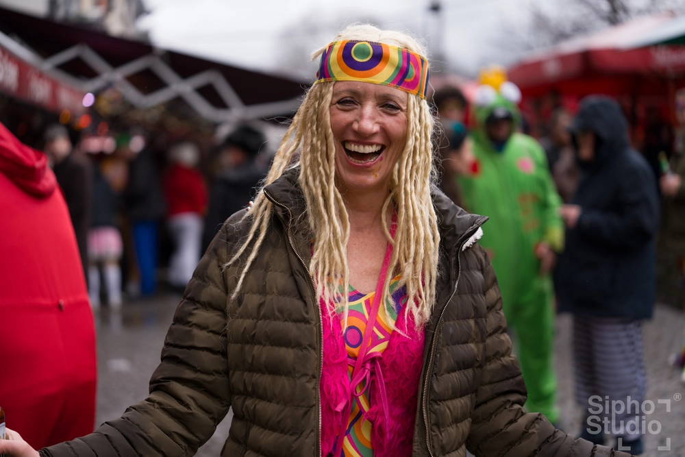 Sipho Studio - Drone visuel - Lachende Frau mit buntem Stirnband, Dreadlocks-Perücke und 70er-Outfit beim Köln Karneval 2025.