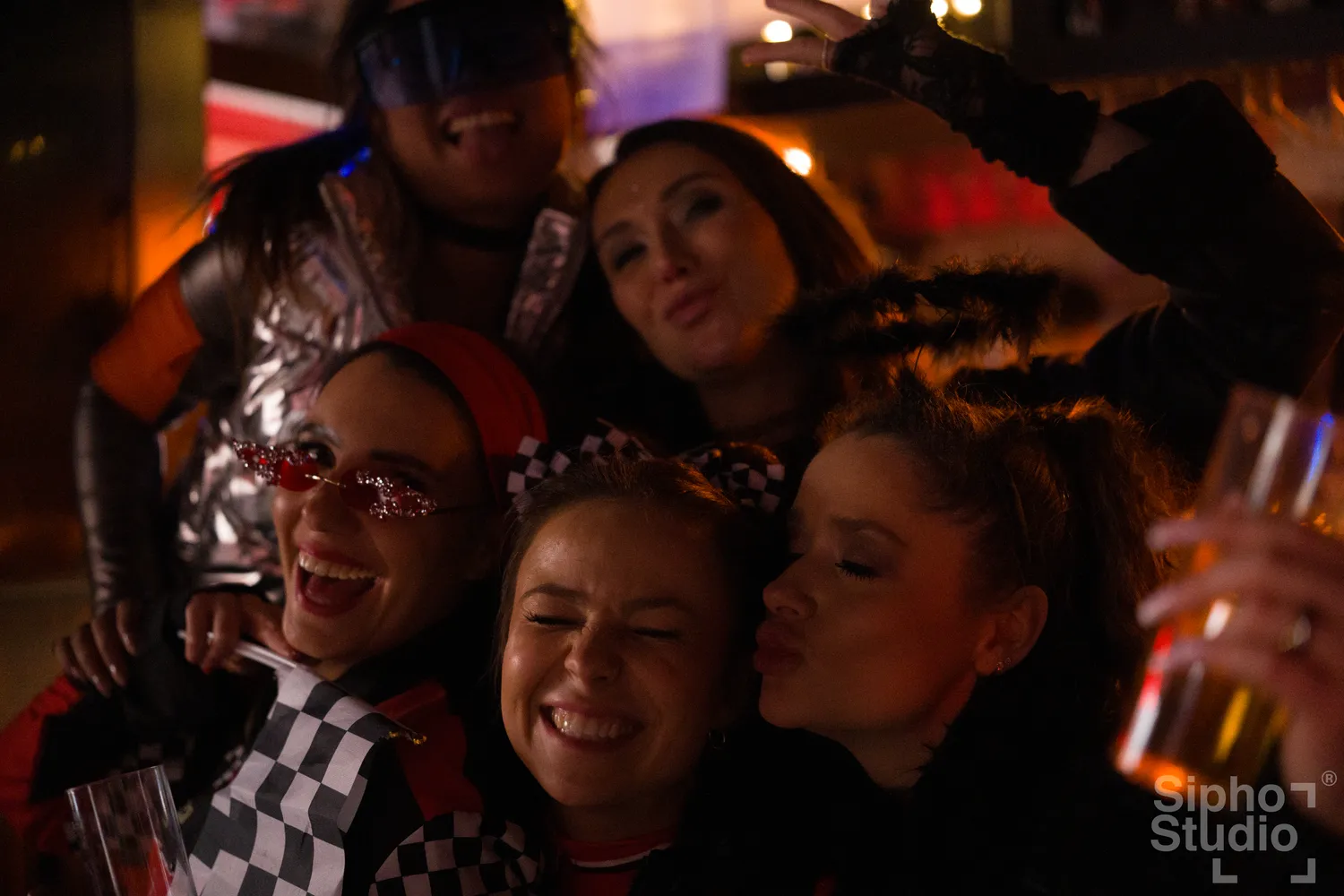 Sipho Studio - Drone visuel - Eine Gruppe lächelnder und posierender junger Frauen, die eng zusammen auf einer schwach beleuchteten Kostümparty feiern.