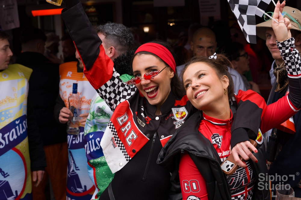 Sipho Studio - Drone visuel - Zwei lachende Frauen in rot-schwarzen Outfits und Karo-Jacken feiern in einer Menschenmenge