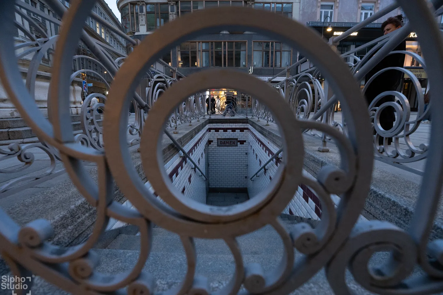 Sipho Studio - Drone visuel - Blick durch ein verziertes Metallgeländer auf eine unterirdische Treppe mit beleuchtetem Eingang.