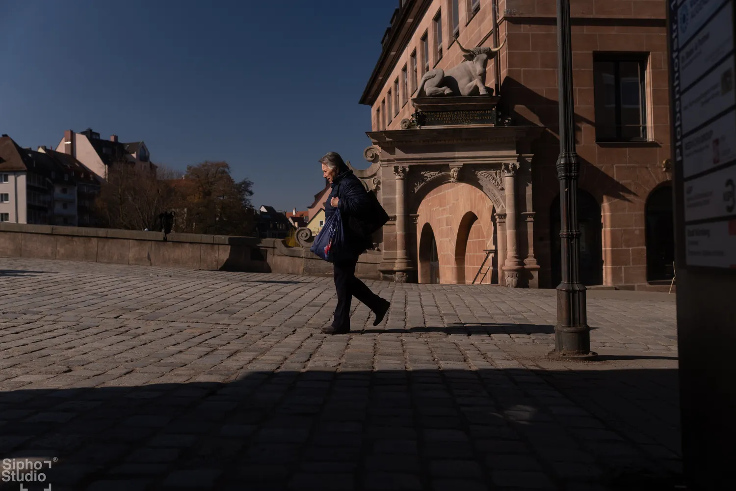 Sipho Studio - Drone visuel - Frau mit Einkaufstüte läuft über einen sonnigen Kopfsteinpflasterplatz vor einem historischen Sandsteingebäude.