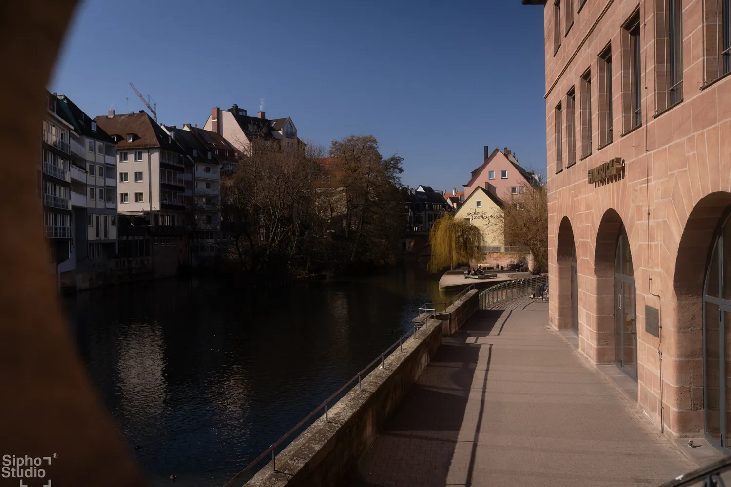 Sipho Studio - Drone visuel - Ruhiger Flusslauf mit Häusern und kahlen Bäumen am Ufer unter klarem blauem Himmel.
