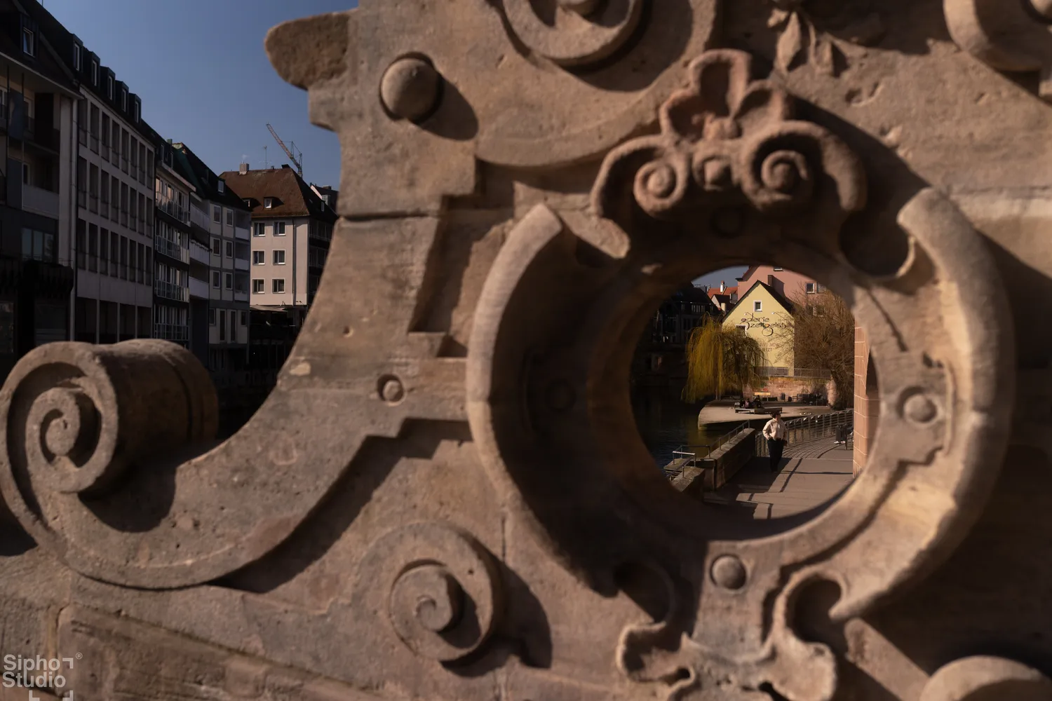 Sipho Studio - Drone visuel - Blick durch eine verzierte Steinöffnung auf einen Fluss mit Häusern und Bäumen im Sonnenlicht.