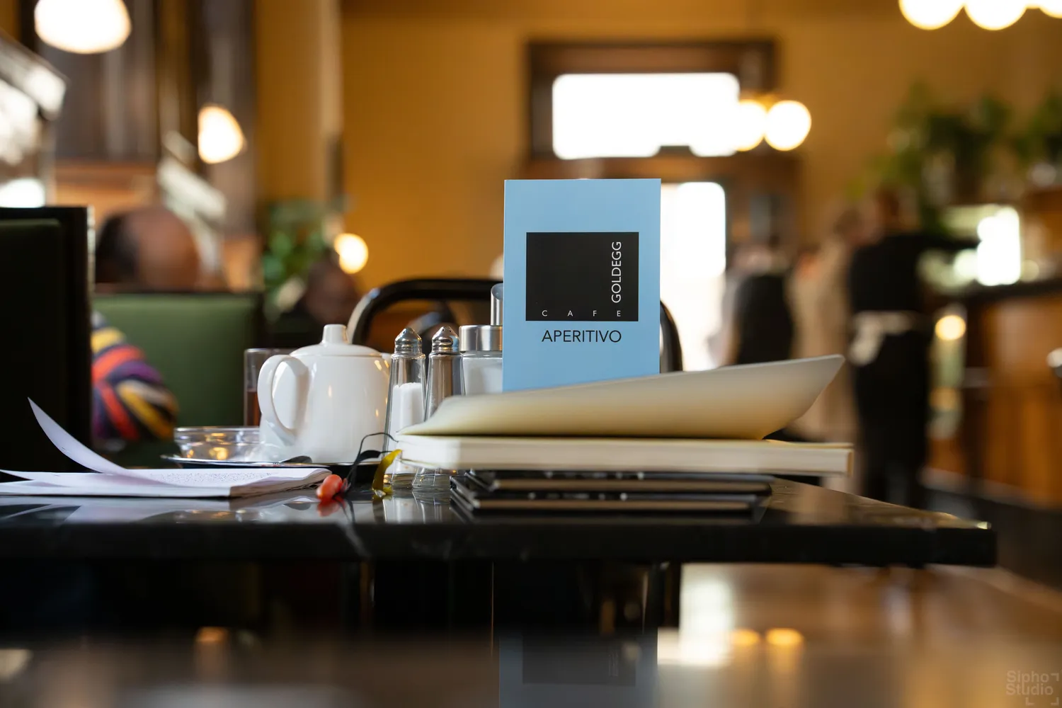 Sipho Studio - Drone visuel - Tisch in einem Café mit hellblauer "APERITIVO"-Menükarte, Teekanne und Salzstreuern im warmen Licht.