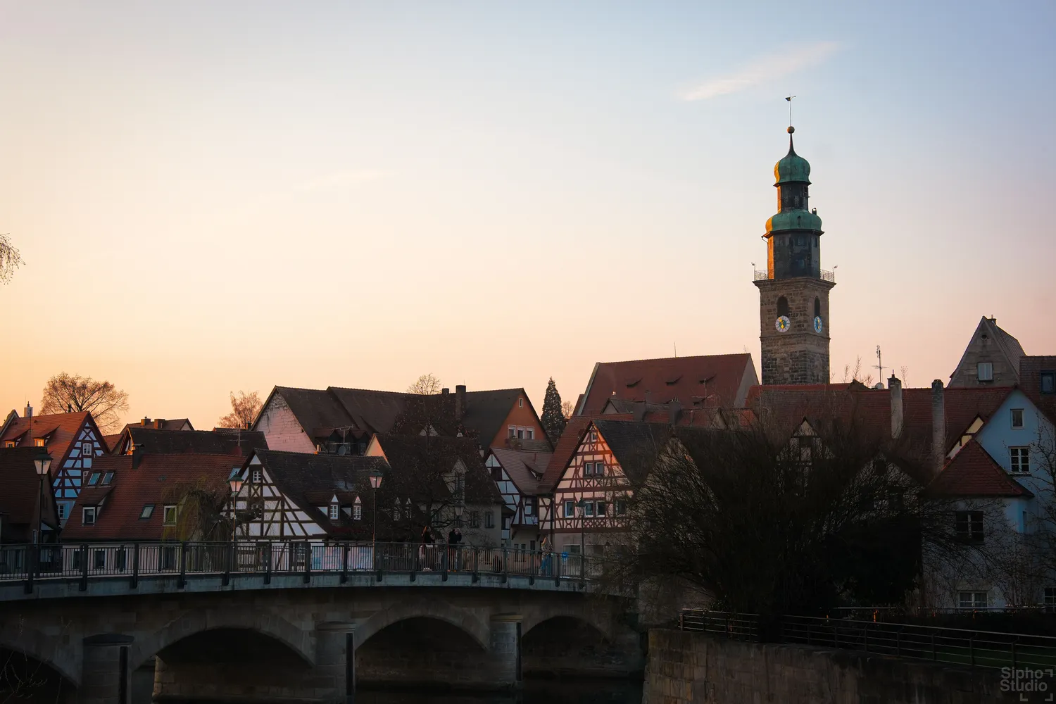 Sipho Studio - Drone visuel - Fachwerkhäuser und Kirchturm mit grüner Kuppel ragen hinter einer Steinbrücke im Abendlicht auf.