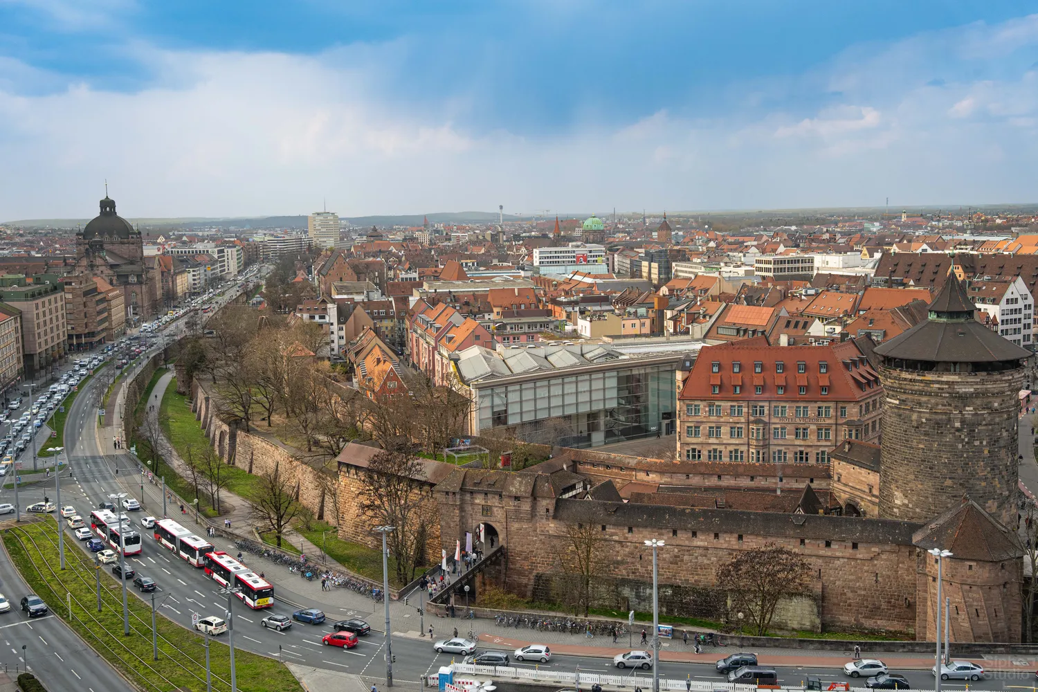 Sipho Studio - Drone visuel - Blick über eine Altstadt mit roten Dächern, Stadtmauer und modernem Glasgebäude unter blauem Himmel.