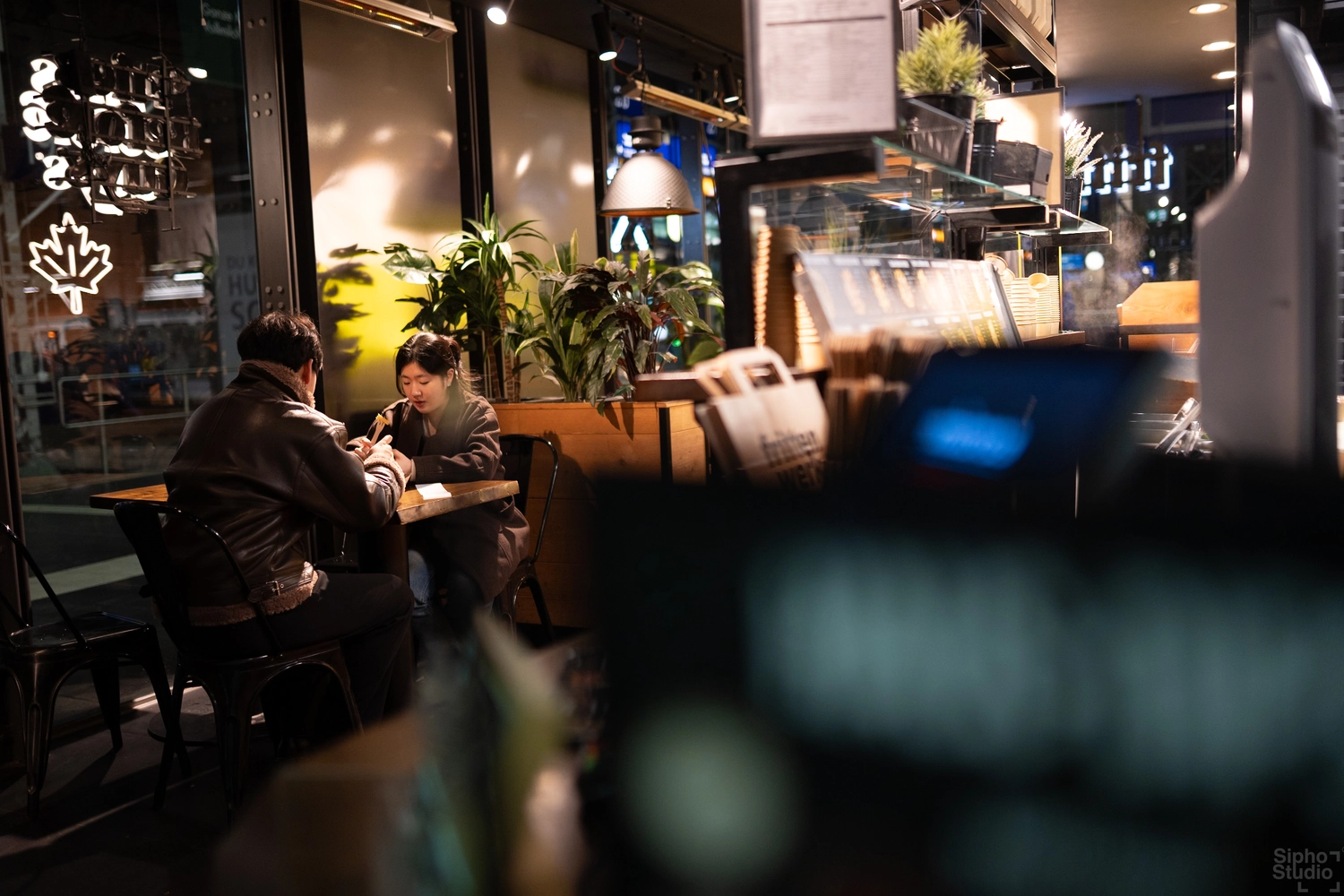 Sipho Studio - Drone visuel - Zwei Personen sitzen an einem Tisch in einem gemütlichen Café mit Pflanzen und Vitrine im Hintergrund.